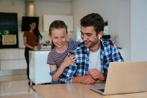 Work from home. Daughter interrupting her father while he is having a business online conversation on his laptop while sitting in the modern living room. photo