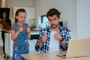Work from home. Daughter interrupting her father while he is having a business online conversation on his laptop while sitting in the modern living room. photo