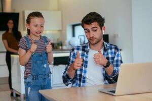 Work from home. Daughter interrupting her father while he is having a business online conversation on his laptop while sitting in the modern living room. photo