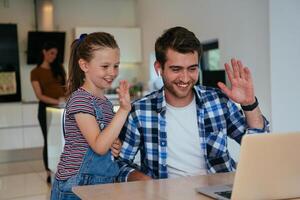 Work from home. Daughter interrupting her father while he is having a business online conversation on his laptop while sitting in the modern living room. photo