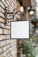 A blank rectangular signboard with a black metal frame hangs from an ornate wrought iron bracket on a vintage brick wall. The softly lit street adds a nostalgic atmosphere photo