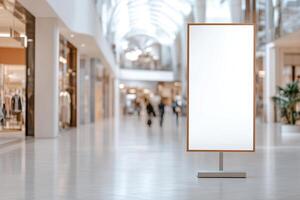 Inside a bright shopping mall, a blank vertical billboard stands ready for use. Stylish storefronts line the walkway, with blurred shoppers exploring various retail options nearby photo