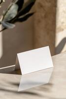 A blank business card stands upright against a neutral-toned wall, slightly leaning. The soft reflections and blurred background create an aesthetic, upscale ambiance ideal for luxury branding photo