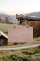 A blank wooden signboard hangs from a wooden post in a serene rural setting. The softly blurred background features rolling hills, a barn, and a dirt road, providing a tranquil atmosphere photo