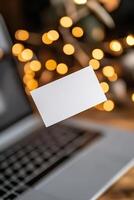 A blank business card is suspended above a laptop, illuminated by soft lighting. The background showcases a modern workspace with a sleek design, perfect for startups and digital branding photo
