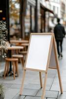 A blank A-frame sandwich board stands prominently on a busy sidewalk in front of a trendy cafe. The scene features wooden tables and people strolling by, creating a lively atmosphere photo