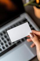 In a sleek, high-tech workspace, a blank business card hovers just above a laptop keyboard, illuminated by warm lighting. The background offers a stylish, innovative atmosphere perfect for startups photo