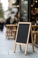 Busy sidewalk features a blank A-frame sandwich board outside a trendy cafe. The background includes wooden tables, a glass storefront, and people strolling by on a sunny day photo
