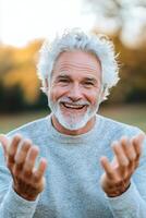 A senior man with white hair and a big smile playfully throws his hands up as if in shock. He enjoys a joyful moment in a sunny outdoor park setting, surrounded by warmth and nature photo