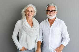 A cheerful senior couple stands side by side, holding hands and smiling at the camera. They wear modern, casual outfits against a soft backdrop, embodying warmth and connection photo