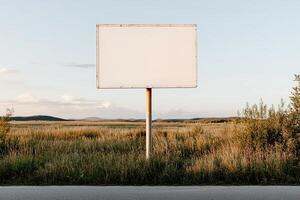 A large blank billboard is situated on a tranquil country road, embraced by expansive green fields and a serene blue sky, making it ideal for minimal branding opportunities photo