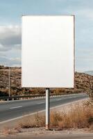 Blank signboard mounted on a sturdy pole stands beside a busy highway. Moving vehicles pass while the landscape features open spaces and a partly cloudy sky, ideal for ads photo