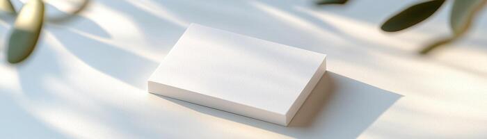 A blank business card rests on a smooth white surface, softly lit by natural light that creates gentle shadows. The background is blurred, highlighting the card's clean design and professional appeal photo
