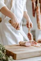 A butcher dressed in a white apron is meticulously cutting fresh meat on a wooden block. The rustic shop features hanging cured meats, creating a classic ambiance photo