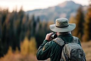 A park ranger in a green uniform stands on a beautiful hiking trail, using binoculars to observe wildlife amidst tall pine trees and a stunning mountain backdrop photo