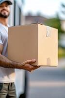 A professional mover in uniform is carrying a labeled cardboard box while standing beside a moving truck. The background features softly blurred suburban homes, creating a friendly atmosphere photo