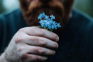 A rugged bearded man stands in soft sunlight, holding a small bouquet of forget-me-nots. The gentle light highlights his hands, symbolizing a poignant connection to nature photo
