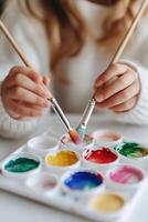 Hands of a child joyfully dip brushes into bright watercolor paints, exploring creativity and color at a table. Soft focus enhances the artistic atmosphere, inspiring early education photo
