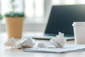 A chaotic home office desk filled with crumpled papers and an overflowing trash can, coffee stains mark important documents while a blurred laptop adds to the stressed atmosphere photo