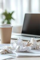 The desk shows a chaotic home office atmosphere with crumpled papers scattered around, an overflowing trash can, and coffee stains. A blurred laptop hints at ongoing work photo