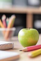 A neat arrangement of colorful school supplies, featuring a shiny green apple, sits on a wooden desk. The softly blurred classroom background creates an inviting educational setting photo