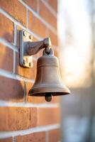Soft sunlight highlights a traditional school bell mounted on a brick wall, offering a nostalgic symbol for announcements in an empty schoolyard. The blurred background enhances the focus on the bell photo