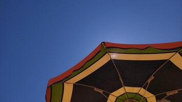 Vibrant beach umbrella casting shadow on sandy shoreline, distant airplane soaring across cloudless azure horizon photo