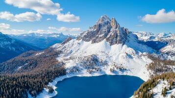 A stunning aerial view of a snow-covered mountain range surrounding a calm blue lake, under a clear blue sky dotted with fluffy clouds. photo