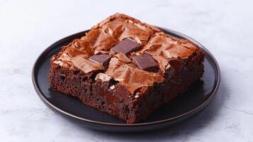 Rich brownie with chocolate chunks concept. A rich, fudgy chocolate brownie topped with chunks of chocolate, served on a black plate against a light background. photo