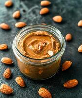 Plant-based diets Food concept. A jar of creamy almond butter sits on a dark surface, surrounded by scattered almonds, showcasing its rich texture and natural ingredients. photo