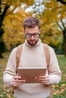 Personalized learning for education concept. A man with glasses reads a tablet while standing in a park adorned with autumn foliage, wearing a cozy sweater and a backpack. photo