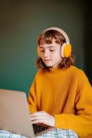 Special needs for Education concept. A young girl wearing headphones types on a laptop, showcasing a cozy atmosphere with a colorful sweater and a green backdrop. photo