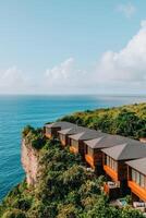Adventure and Eco-tourism for travel concept. Cliffside resort overlooks the ocean, featuring modern wooden accommodations surrounded by lush greenery and a bright blue sky. photo