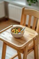 Parenting resources for family concept. Wooden high chair holds a bowl of colorful mixed vegetables, suggesting a cozy dining setting for a child. photo