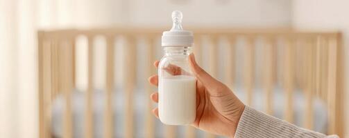 paternidad recursos para familia concepto. mano sostiene un bebé botella lleno con leche, conjunto en contra un enfoque suave antecedentes de un guardería con un cuna. foto