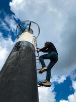 A determined man is climbing a tall, sturdy pole using a solid ladder, focused on reaching the very top safely as he steadily ascends with persistence and resolve to achieve his goal photo