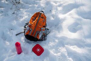 A bright orange backpack is resting in the softly falling snow next to a vibrant red first aid kit, which creates an eyecatching contrast against the stunningly beautiful wintry landscape photo