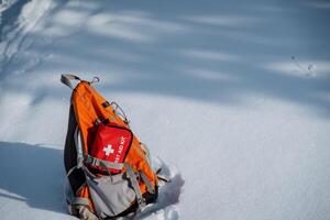 A backpack, which contains a first aid kit for emergencies, is currently resting in the snow, surrounded by a wintry landscape, creating a stark contrast against the white backdrop photo