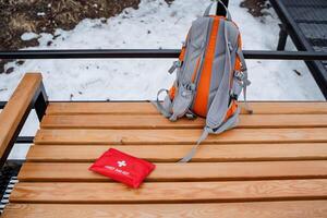There is a backpack that is resting on a wooden bench that is positioned right beside a first aid kit, creating a convenient resting spot for outdoor enthusiasts or hikers photo