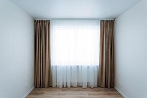The room is completely empty, featuring a large window dressed with brown curtains that gently sway in the soft breeze, providing a sense of openness and light to the space photo