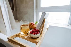 A beautifully crafted wooden bathtub tray is adorned with fresh, plump raspberries, a flaky croissant, a glass of fine wine, and an engaging book to read during a relaxing bath photo
