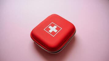 On a soft pink surface, you will find a vibrant red first aid kit featuring a prominent white cross design on its exterior, which signifies its purpose and importance in medical care photo