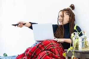 Woman with dreadlocks working with a laptop at home and with a remote control in hand photo