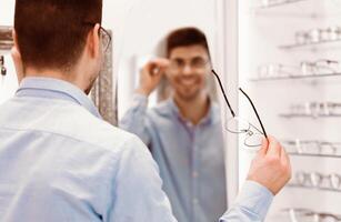 A man stands in front of a mirror, holding a pair of glasses and smiling. He is in an eyewear store, surrounded by various frames, creating a cheerful atmosphere. photo