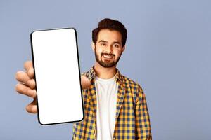 Smiling Indian guy showing smartphone with mockup for mobile app design on screen, violet background photo