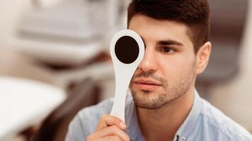 A young man focuses intently while holding an occluder over one eye during a vision examination in a modern eye care clinic, highlighting the importance of visual health. photo