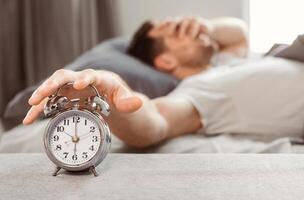 Bad Sleep. Sleepy Man Turning Off Alarm-Clock Lying Waking Up In Bed At Home. Shallow Depth, Selective Focus photo