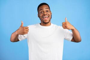 Black Millennial Guy Gesturing Thumbs Up With Both Hands Smiling To Camera Standing Over Blue Background In Studio photo