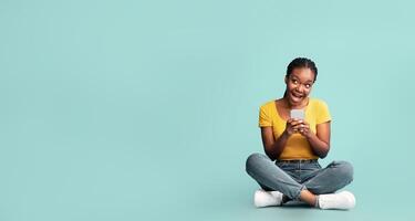 A happy young woman sits cross-legged on the floor, holding her smartphone and smiling cheerfully, surrounded by a calming green background that enhances her joyful expression. photo