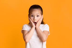 Wow. Portrait of surprised and shocked asian girl touching cheeks with open mouth in amazement isolated over yellow studio background. Child saying omg or oops, no way, looking at camera photo